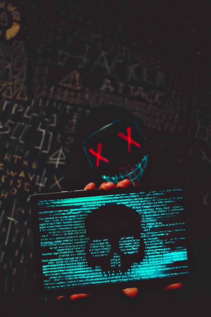 Mysterious hacker with neon mask holding a tablet displaying digital skull against a dark backdrop.